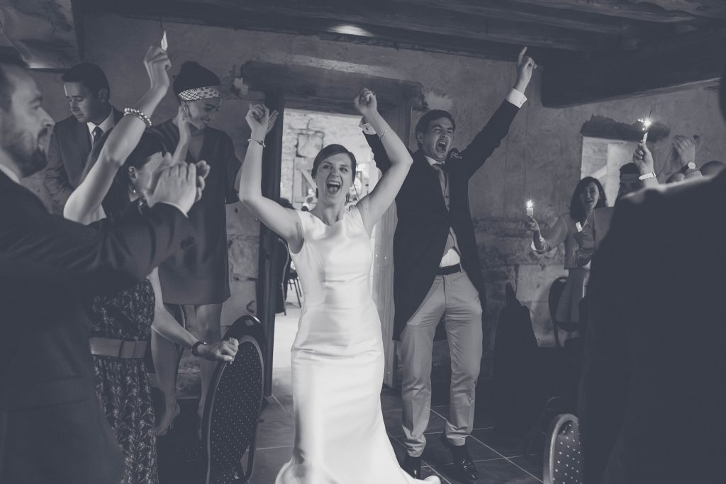 La mariée bras levés à son entrée dans la salle de réception, invités applaudissant aux cierges magiques, noir et blanc