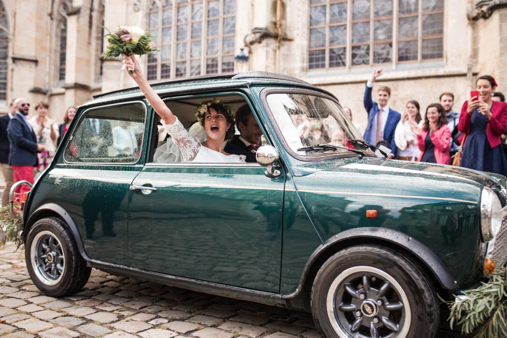 Sortie de mariage à la Cathédrale de Nevers – La mariée souriante dans une voiture rétro sous le soleil après la pluie.